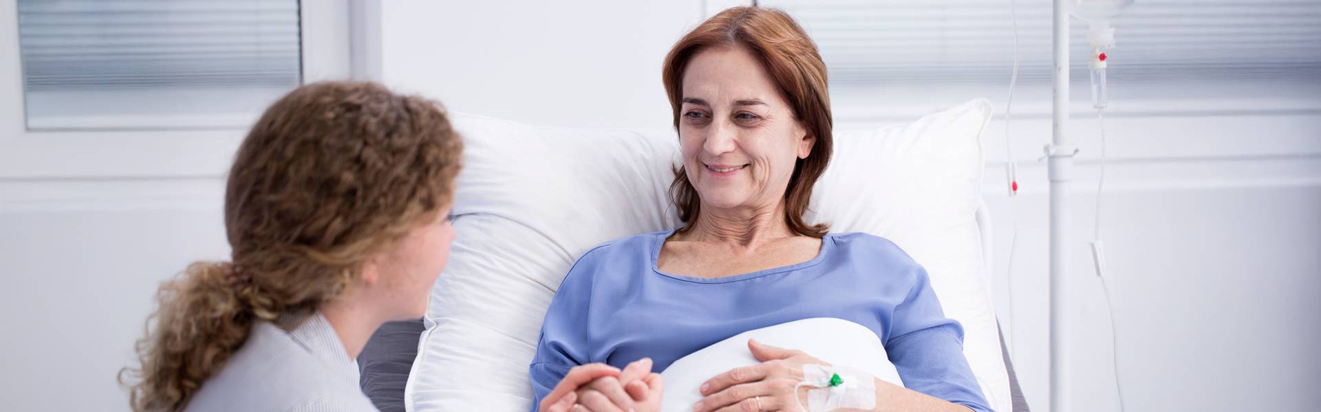 Frau mit langen Haaren, Zopf sitze am Bett einer Patientin und hält ihr die Hand